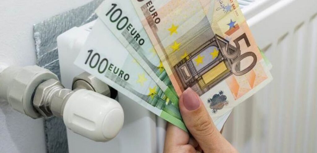 Επίδομα θέρμανσης: Ένα χέρι κρατά 2 χαρτονομίσματα των 100 ευρώ και ένα των 50 μπροστά από ένα σώμα καλοριφέρ