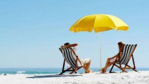 δύο άνθρωποι κάθονται σε ξαπλώστρες μπροστά στη παραλία κάτω από μια κίτρινη ομπρέλα