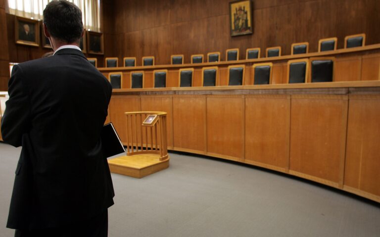 Δικαστική αίθουσα, δεξιά οι άδειες καρέκλες των δικαστικών, αριστερά ένας κύριος με κουστούμι και πλάτη προς το φακό