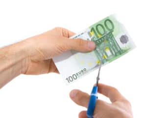 χέρι κρατά χαρτονόμισμα των 100 ευρώ και ένα άλλο χέρι κρατά ψαλίδι και επιχειρεί να κόψει το χαρτονόμισμα