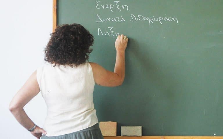 Καθηγήτρια με άσπρη μπλούζα; γράφει με κιμωλία πάνω σε πράσινο πίνακα
