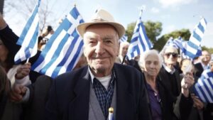 ηλικιωμένοι άνδρες και γυναίκες σε συγκέντρωση κρατώντας ελληνικές σημαίες