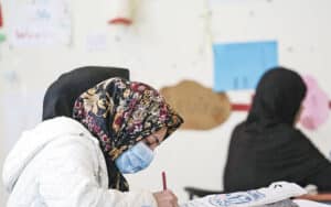 γυναίκα με μάσκα στο πρόσωπο και μαντίλι στο κεφάλι γράφει σε εξεταστικό κέντρο