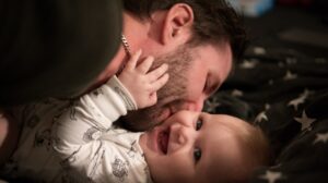 άντρας φιλάει ένα μωράκι στο μάγουλο το οποίο είναι ξαπλωμένο σε ένα κρεβάτι