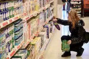 μια γυναίκα σε σούπερ μάρκετ κρατά στα χέρια της δύο προϊόντα και ταυτόχρονα ελέγχει τη τιμή που αναγράφεται στο ράφι 