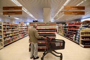 ένας κύριος κρατώντας ένα άδειο καροτσάκι του super market βρίσκεται μπροστά σε ένα διάδρομο με προϊόντα και εξετάζει τις τιμές 