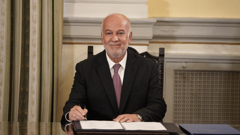 Ο υπουργός Δικαιοσύνης Γιώργος Φλωρίδης με σκουρόσρωμο κουστούμι και μωβ γραβάτα υπογράφει